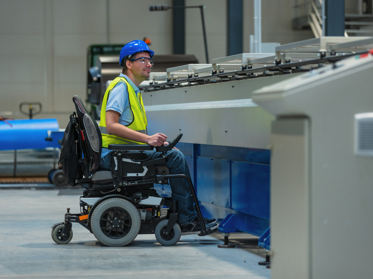 A man in a wheel chair wearing a hardhat and a safety vest inspecting a large industrial machine in a factory