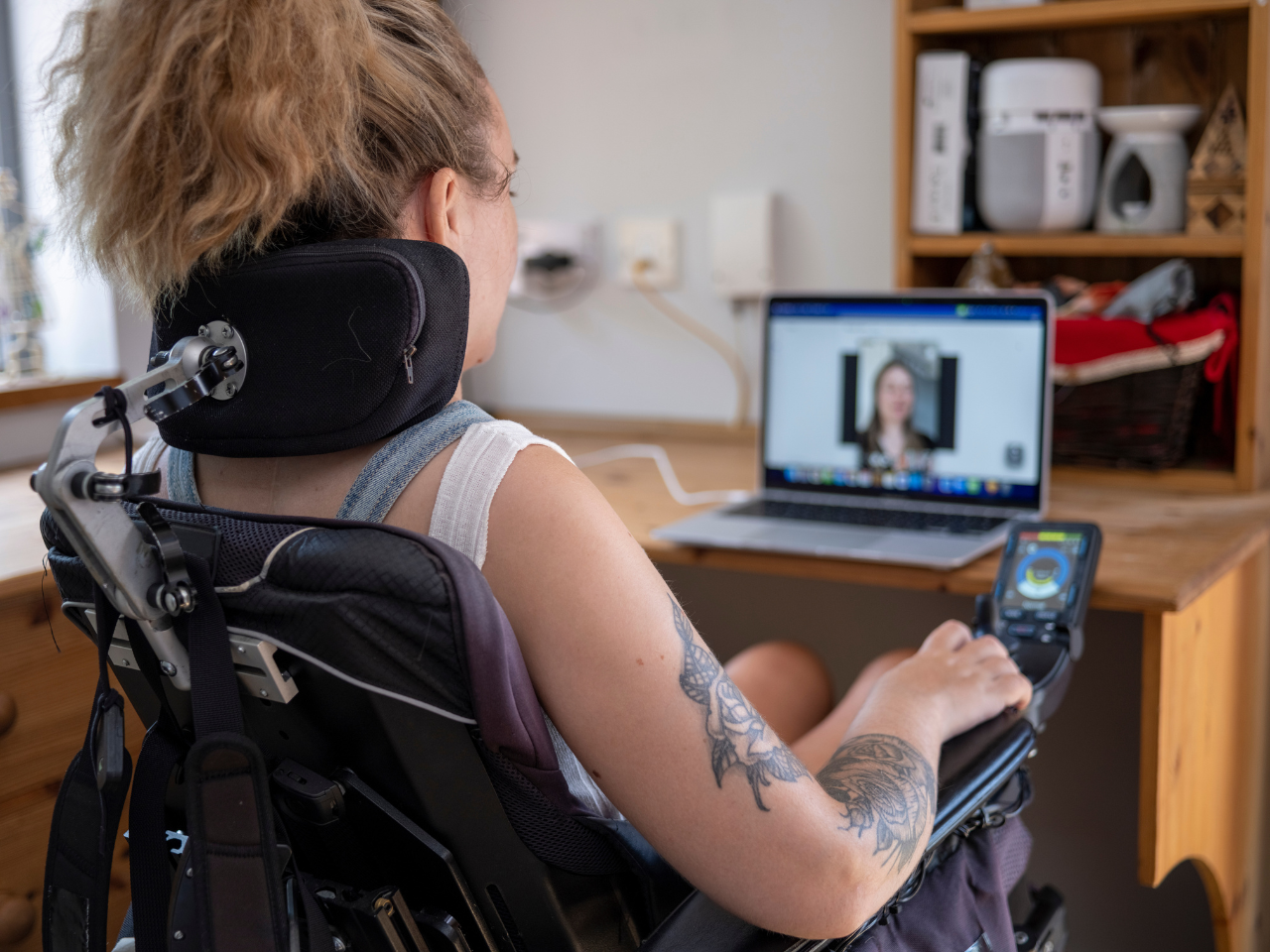 A young woman in a wheelchair utilizing assistive technology to engage in a video conference call from a dorm room